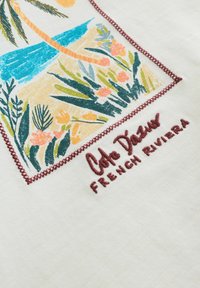 Impression colorée de plantes et fleurs tropicales bordée de rouge avec le texte brodé "Côte d'Azur FRENCH RIVIERA" sur tissu blanc.