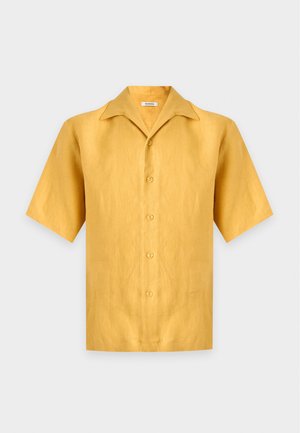 Homme portant une chemise à manches courtes jaune moutarde avec boutons, pantalon marron foncé, baskets blanches, debout les mains dans les poches devant un fond uni.