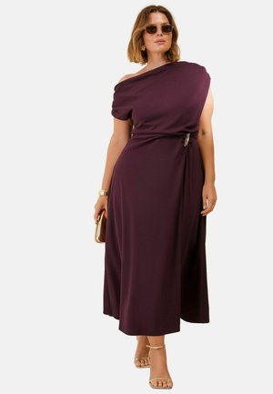 Woman wearing one-shoulder dark purple midi dress with a side belt, sunglasses, gold bracelet, clutch, and strappy high-heeled sandals.