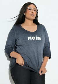 Granatowa koszula z rękawem do połowy ramienia i dekoltem w serek. Na niej biały nadruk z napisem "MOIN" oraz wzór kotwicy. Wykonana z miękkiego, fakturowanego materiału.