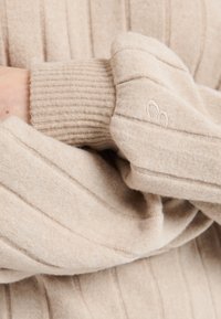 Beżowy sweter dziany z pionową prążkowaną fakturą. Posiada mankiet na rękawie oraz subtelny haft przy nadgarstku. Wykonany z miękkiej mieszanki wełny.