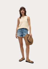 Beige gebreide tanktop, versleten denimshorts, een bruine strooien hoed met brede rand en bruine instapsandalen met studs. Casual zomerse outfit.
