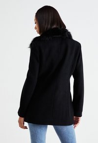 Čierny kabát s umelou kožušinou na golieri, strihaný dizajn, dlhé rukávy, hladká textúra a štruktúrovaný strih, oblečený cez svetlo modré džínsy.