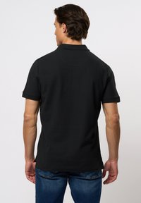 Zwarte poloshirt van een gestructureerde stof, korte mouwen en een klassieke kraag. Heeft een subtiele naaddetail op de rug. Gekomd met blauwe jeans.