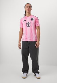 Rosa Sportjersey mit schwarzen Akzenten, featuring ein zentrales Logo und einem gestreiften Design. Getragen mit dunklen weiten Hosen und weißen Sneakers.