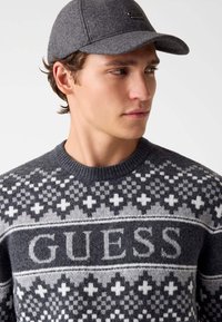 Pull en laine gris avec des motifs géométriques blancs et noirs, arborant un logo "GUESS" bien en vue. Casquette en feutre gris avec une visière rigide.