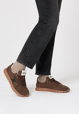 Sneakers marroni con una tomaia texturizzata, accenti neri e suola in gomma marrone. Abbinati a calzini grigi e jeans scuri.