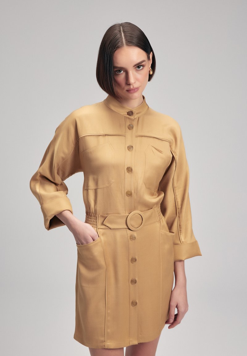 Abito camicia belted color beige con colletto, chiusura a bottoni e due tasche sul petto. Texture morbida con una finitura leggermente lucida.