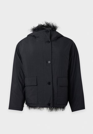 Femme portant une veste noire doublée en fausse fourrure, un pull beige, une longue jupe en cuir noire et des bottines noires à talons, debout sur un fond blanc.