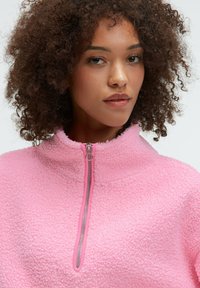 Fleece-Pullover in leuchtendem Pink mit hohem Kragen und halbem Reißverschluss, der eine strukturierte, flauschige Oberfläche und einen kontrastierenden Reißverschluss aufweist.