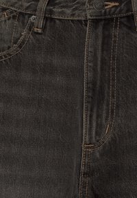 Gros plan sur un jean en denim noir montrant la poche avant, la boucle de ceinture, le bouton et les détails de couture orange.