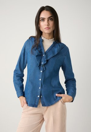 Dunkelblaue Jeanshemd mit Rüschensaum und Knöpfen auf der Vorderseite, kombiniert mit hellbeigen Cordhosen. Insgesamt glatte Textur.