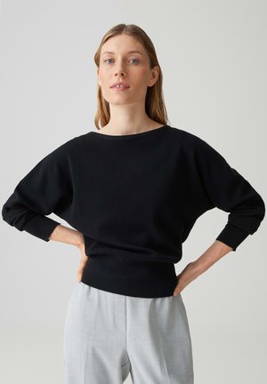 Frau mit langen blonden Haaren, die einen schwarzen Pullover und hellgraue Hose trägt, steht mit den Händen in der Hüfte vor einem schlichten Hintergrund.