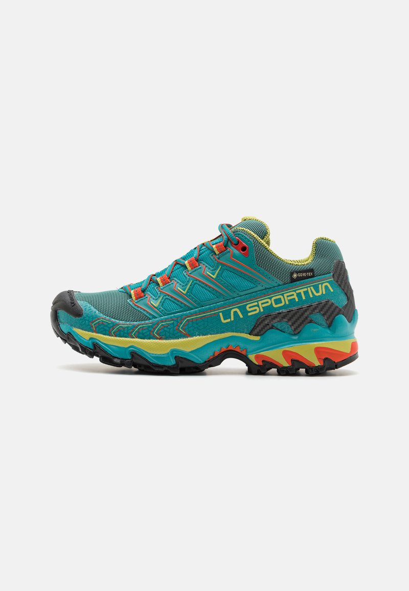 Zapatilla de trail running con parte superior de malla transpirable en turquesa, detalles rojos, puntera de goma negra y suela multicolor texturizada para tracción.