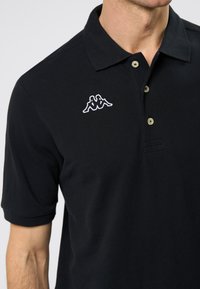 Juodas medvilnės polo marškinėliai su apykakle, trimis mygtukais ir išsiuvinėtu baltu logotipu kairėje krūtinėje. Trumpi rankovės, tekstūruotas audinys.