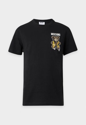 Młody mężczyzna stoi, ubrany w czarną koszulkę Moschino z grafiką misia, czarne spodnie o szerokich nogawkach i czarne sportowe buty, białe tło.