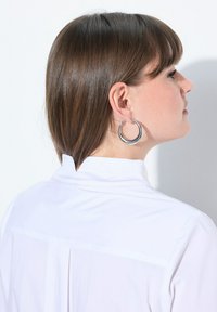 Orecchini a cerchio in argento con una finitura lucida, forma circolare e un leggero splendore, indossati da una persona con una camicia bianca con colletto.