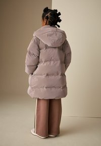 Pufi, világoslila dzseki kapucnival és puha textúrával, látható varrással és laza formával. Barna, bő szárú nadrággal párosítva.