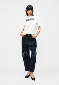 Jeune femme portant un t-shirt blanc Moschino Milano, un jean foncé avec des patchs en forme de cœur aux genoux, et des talons noirs pointus, debout les mains dans les poches.