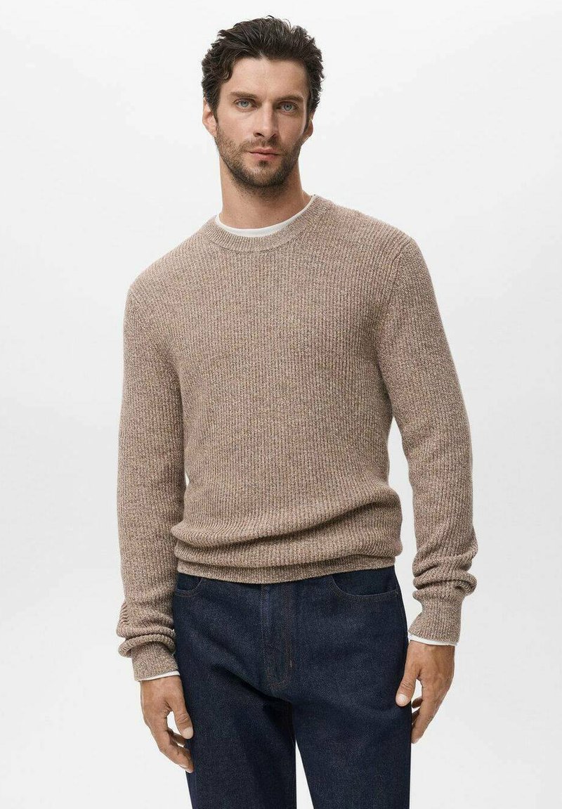 Man in een beige geribbelde trui over een wit shirt, gecombineerd met donkerblauwe jeans, staande tegen een effen achtergrond.