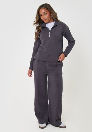 Mujer con camiseta de manga larga gris oscuro con cremallera y pantalones anchos a juego, de pie con la mano en la cadera sobre fondo liso.