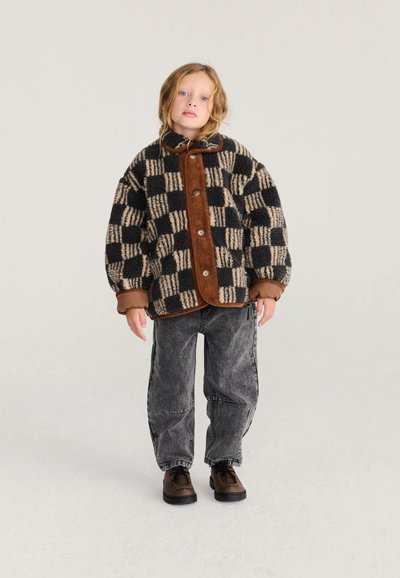 Enfant aux cheveux clairs portant une veste à carreaux noire et beige, un jean gris ample et des chaussures marron, debout sur un fond neutre.