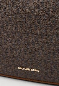Geantă de mână texturată maro cu un model logo și un accent de branding auriu. Material neted, cu un design structurat.