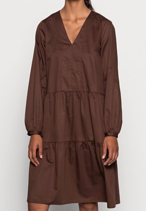 Donna che indossa un vestito marrone largo a maniche lunghe con scollo a V e balze raccolte, in piedi contro uno sfondo chiaro e uniforme.