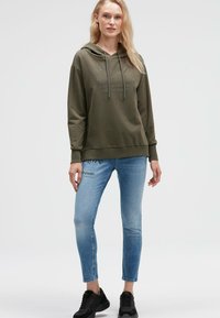Olivgrüner Hoodie mit einer Kordelzugkapuze und einem Front-Text-Design, kombiniert mit hellblauen Slim Jeans mit einem dezenten Druck und schwarzen Sneakers.