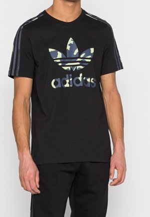 Schwarzes Baumwoll-T-Shirt mit einem camouflagemusterierten Adidas-Logo in Grüntönen und Blautönen. Mit gestreiften Akzenten an den Schultern.