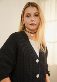 Černý pletený cardigan s výstřihem do V, s velkými perlovými knoflíky a žebrovanou texturou. Nosený přes světle zbarvený top.