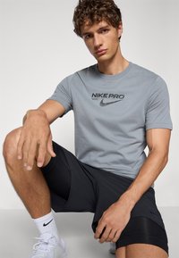 Grå Nike Pro T-skjorte med svart logo, kombinert med svarte treningsshorts. Modell sitter, iført hvite sneakers og ankelhøye sokker.