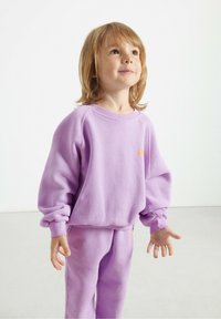 Fioletowy polarowy sweter z rękawami raglanowymi i elastycznymi mankietami, w zestawie z dopasowanymi spodniami. Małe pomarańczowe logo na piersi. Gładka faktura.