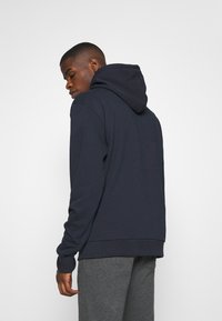 Marineblå hættetrøje med en stor hætte, lange ærmer og ribbede manchetter. Blød tekstur, let løs pasform, iført grå sweatpants.