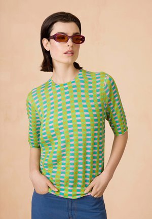 Korte mouwen, textuur top met een groen, blauw en roze dambordpatroon. Gestyled met blauwe broek en zonnebril.