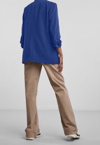 Långärmad, kungligt blå blazer med rak skärning och ryggslits, kombinerad med sandfärgade vida byxor och sportskor med gul accent.