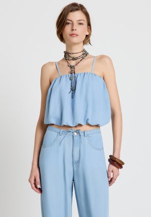 Femme portant un crop top sans manches bleu clair et un pantalon taille haute assorti, accessoirisée avec un collier en couches et des bracelets marron au poignet.