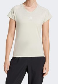 Ljusbeige atletisk t-shirt i fukttransporterande tyg, med V-hals, korta ärmar och en liten vit logotyp på bröstet.