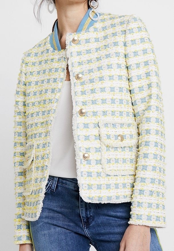 Femme portant une veste à carreaux texturée jaune pastel et bleue avec des boutons dorés, associée à un haut blanc et un jean bleu.