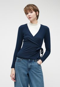 Navy pitkähihainen kietaisupaita, joka on valmistettu pehmeästä kankaasta ja jossa on vyö vyötäröllä, yhdistettynä löysiin vaaleansinisiin farkkuihin, joiden alla on pyöreäkauluksinen valkoinen paita.