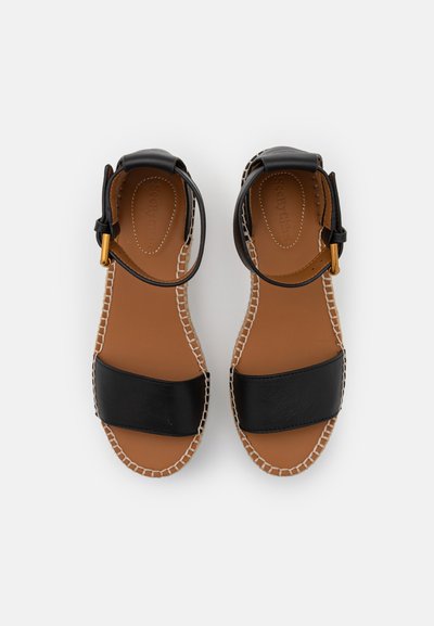 Sorte skinnsandaler med en tykk stropp, ankelspenne og beige såle. Har flettet kantdetaljering og flat hæl.