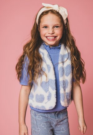 Blauwe fleece vest met witte hartpatronen, gedragen over een blauwe ribbeltop. Gestyled met lichtblauwe jeans met hartpatronen en een lichtkleurige hoofdband.