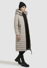 Cappotto puffer lungo beige con motivo a cuscino orizzontale, colletto alto, cerniere frontali, abbinato a pantaloni neri e stivali neri chunky.