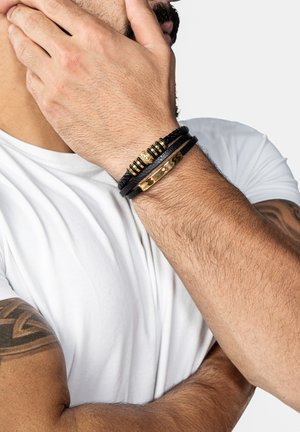 Drei Armbänder an einem Handgelenk: eines aus geflochtenem schwarzem Leder, eines mit goldenen Akzenten und eines minimalistisches schwarzes Band mit einem wellenförmigen Muster.