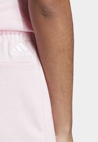 Ljusrosa Adidas mjukisbyxor med elastisk midjeband och sidoficka, med en vit logotyp på vänster höft. Mjuk tygstruktur synlig.