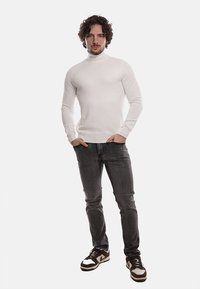 Maglione bianco a collo alto con un motivo testurizzato, abbinato a jeans grigi e sneakers multicolore, creando un outfit casual.