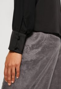 Schwarze Bluse mit einem Puffärmel-Design, kontrastierendem schwarzen Bündchen und zwei Knöpfen. Der Stoff hat eine glatte Textur und wird mit grauen Hosen kombiniert.