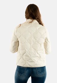 Giacca imbottita crema trapuntata con motivi arrotondati sulla superficie, caratterizzata da un collo alto e una texture morbida, abbinata a jeans blu.