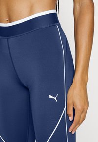Granatowe legginsy sportowe z wysokim stanem, białym akcentem w postaci lampasów i małym białym logo na dolnej części nogawki. Gładki, elastyczny materiał.