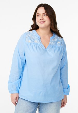 Camicia di cotone azzurro chiaro con scollo a V e ricami floreali sul colletto. Maniche lunghe con polsini elasticizzati e vestibilità ampia.